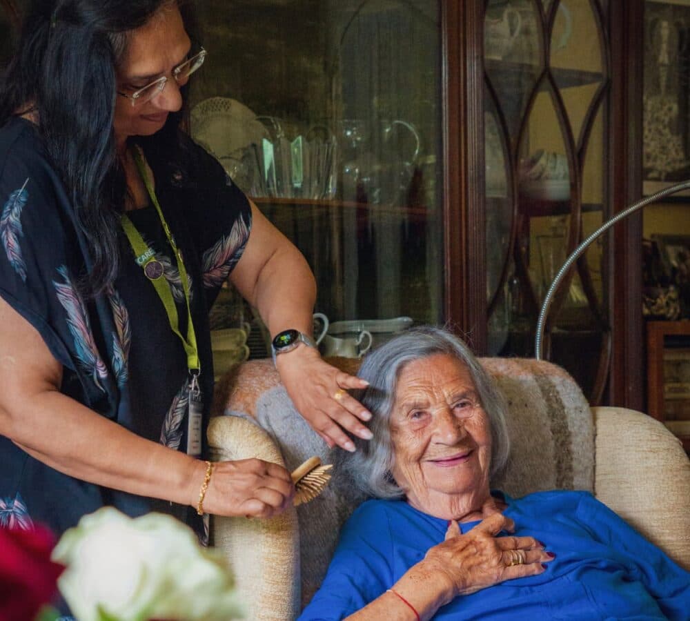 An older female adult sitting on the couch while her hair is being combed by her female carer