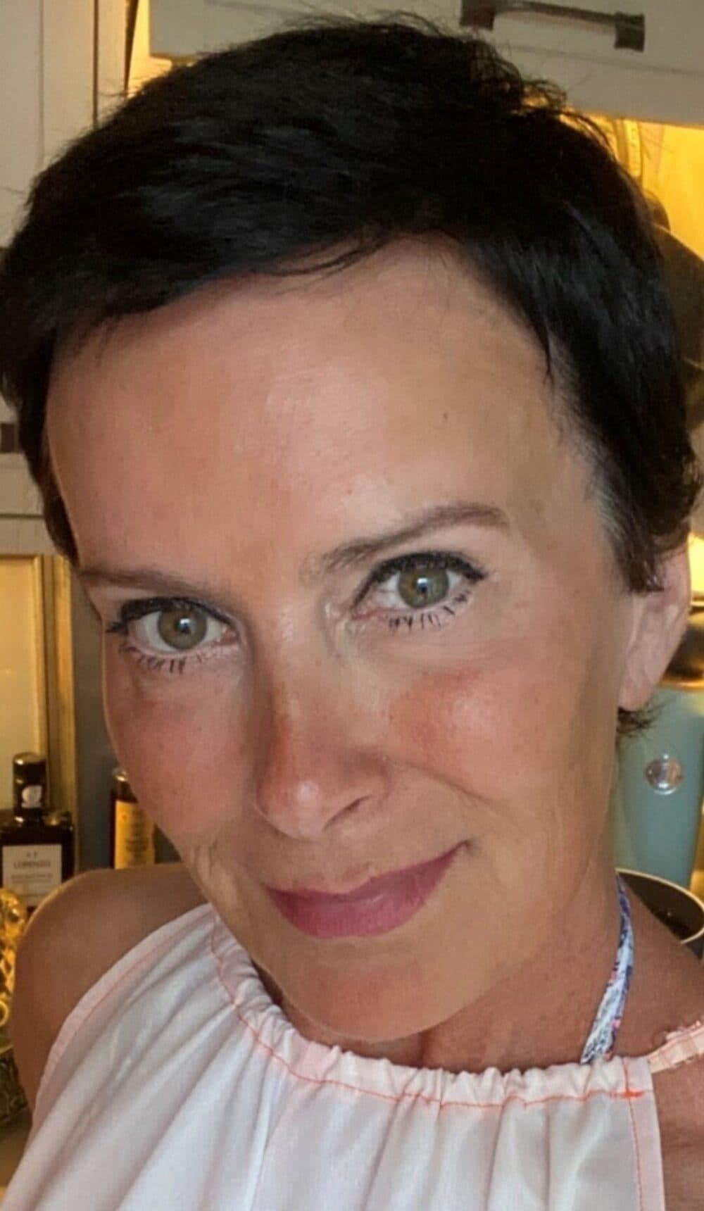 A woman with short dark hair and green eyes smiles softly at the camera in a kitchen setting. - Home Instead