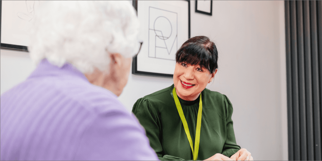 A woman in a green jumper smiles whilst talking to an older woman with white hair and a purple jumper. - Home Instead
