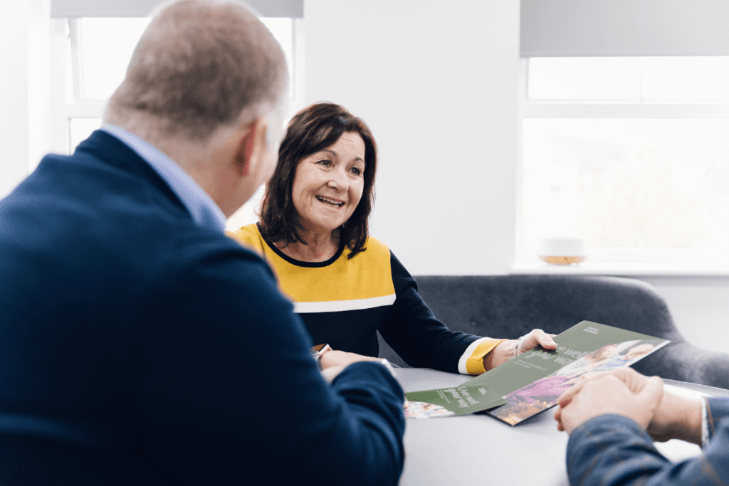 A woman smiling and holding leaflets while talking with two people at a table in a bright office. - Home Instead
