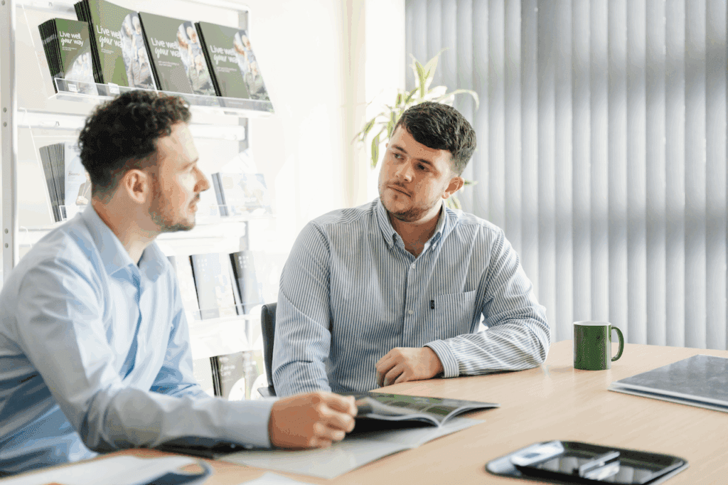 Two men in an office sit at a table, talking with open magazines and a green mug nearby. - Home Instead