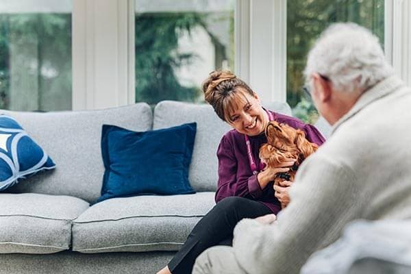 A woman with a small dog smiles at an elderly person in a cozy living room with a grey couch and blue pillows. - Home Instead