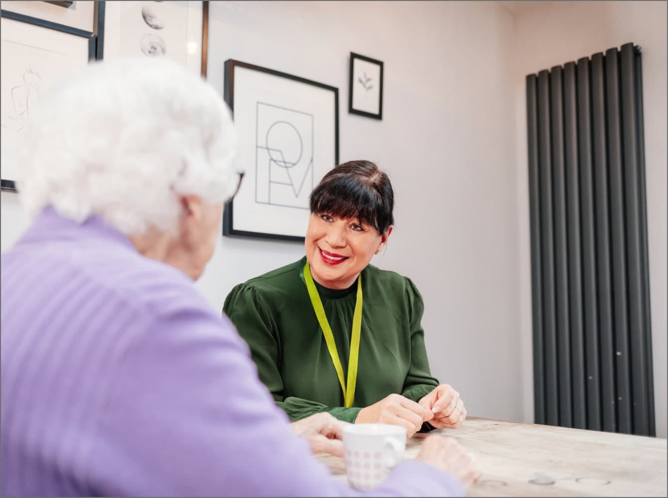 An elderly woman smiles while sitting at a wooden table in a café, with a cup of coffee in front of her. - Home Instead