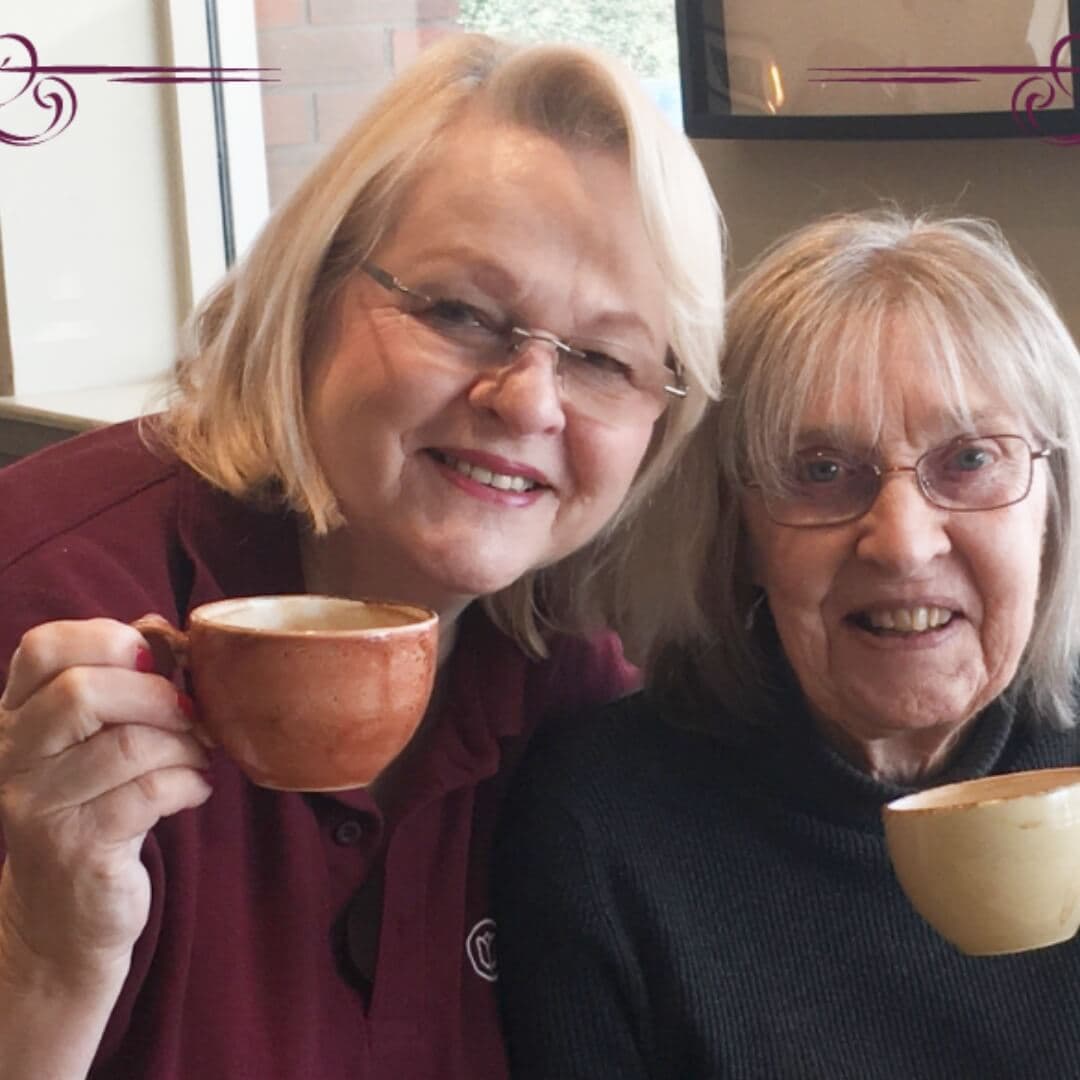 Two smiling women holding coffee cups, seated together in a cozy setting. - Home Instead