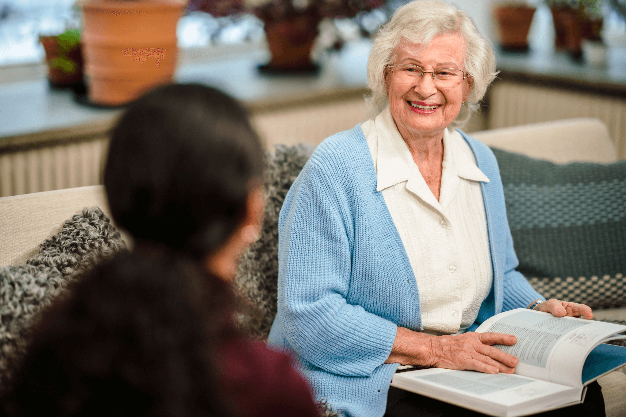 An elderly woman smiling while holding an open book, conversing with another person seated across from her. - Home Instead