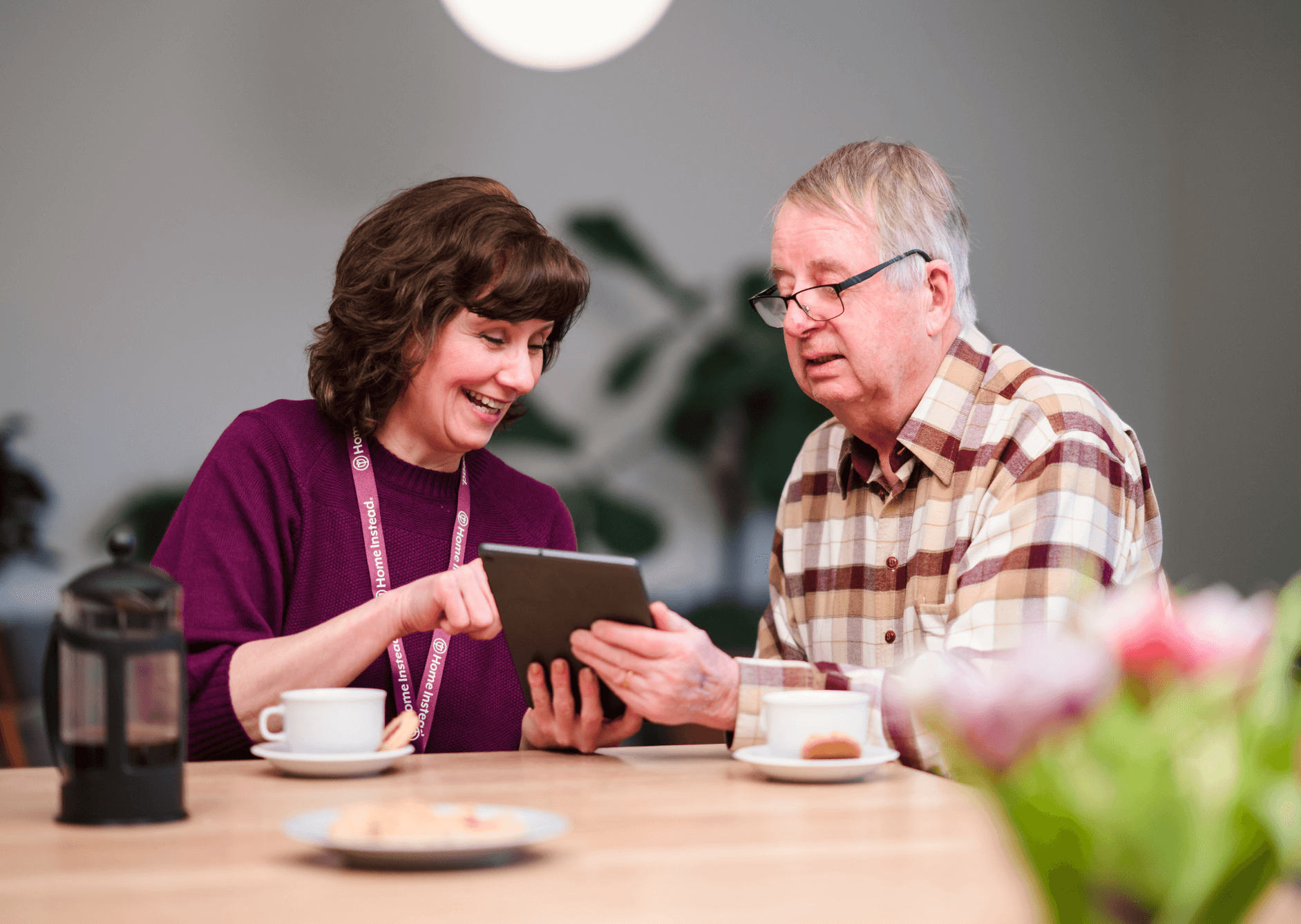 Woman showing an older man something on a tablet at a table with coffee and snacks. They are both smiling. - Home Instead
