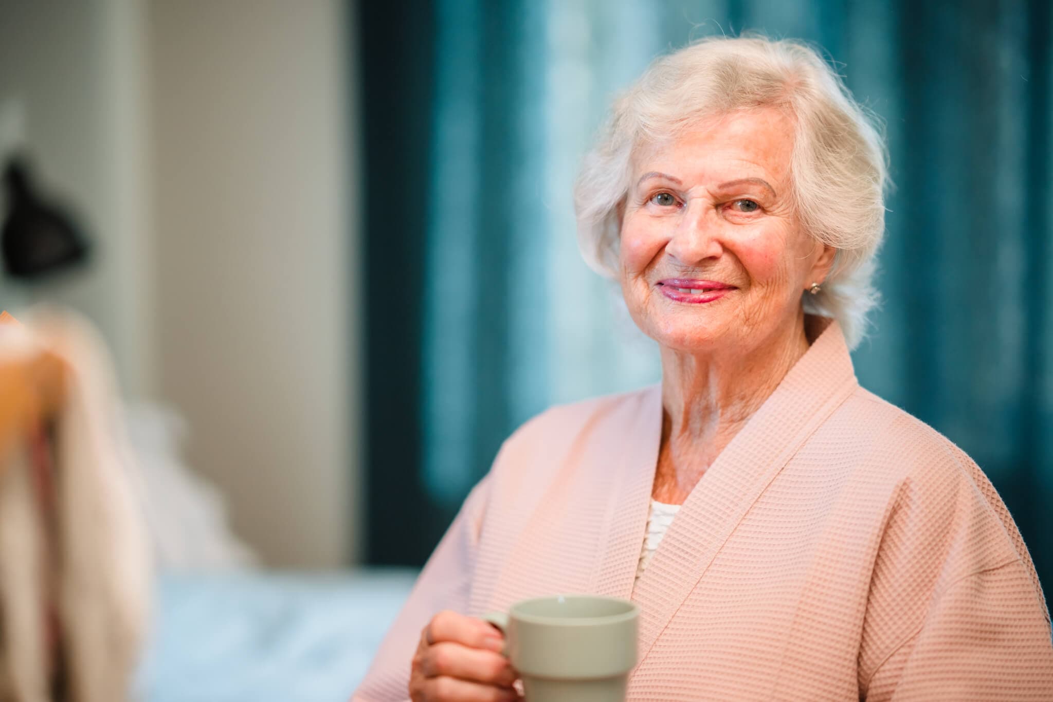 Elderly woman with gray hair smiling and holding a mug while wearing a pink robe, with a blurred background. - Home Instead