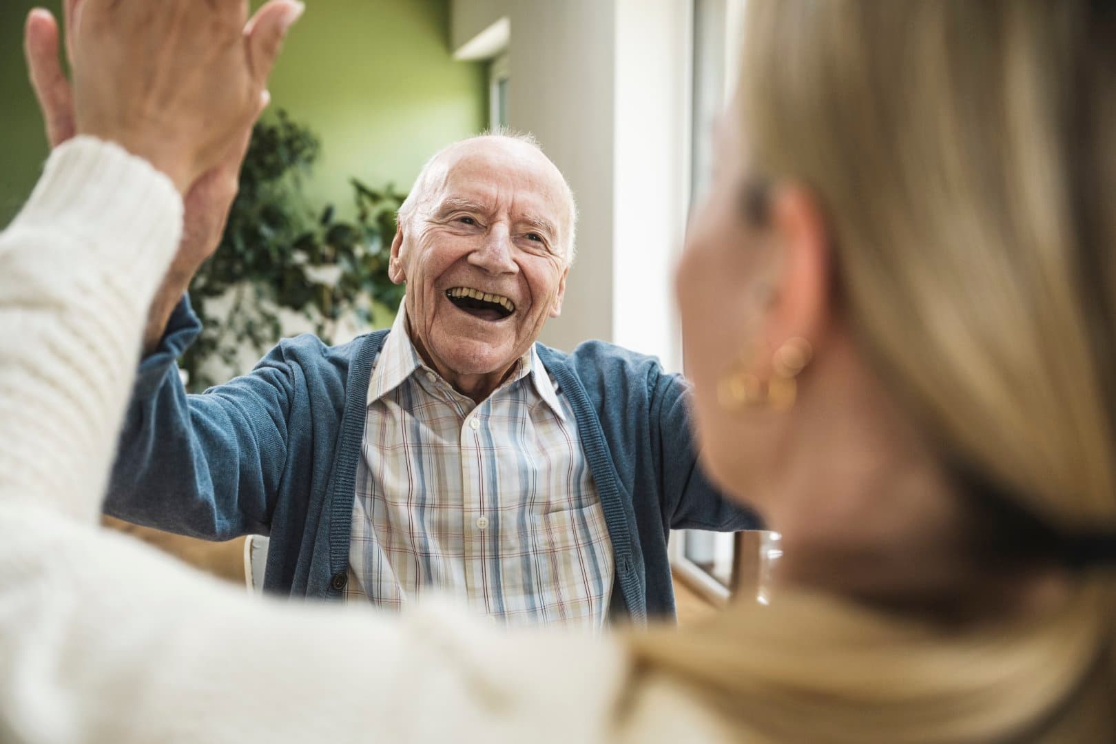 Smiling elderly man in a checked shirt high-fives a woman indoors, both appearing happy and joyful. - Home Instead