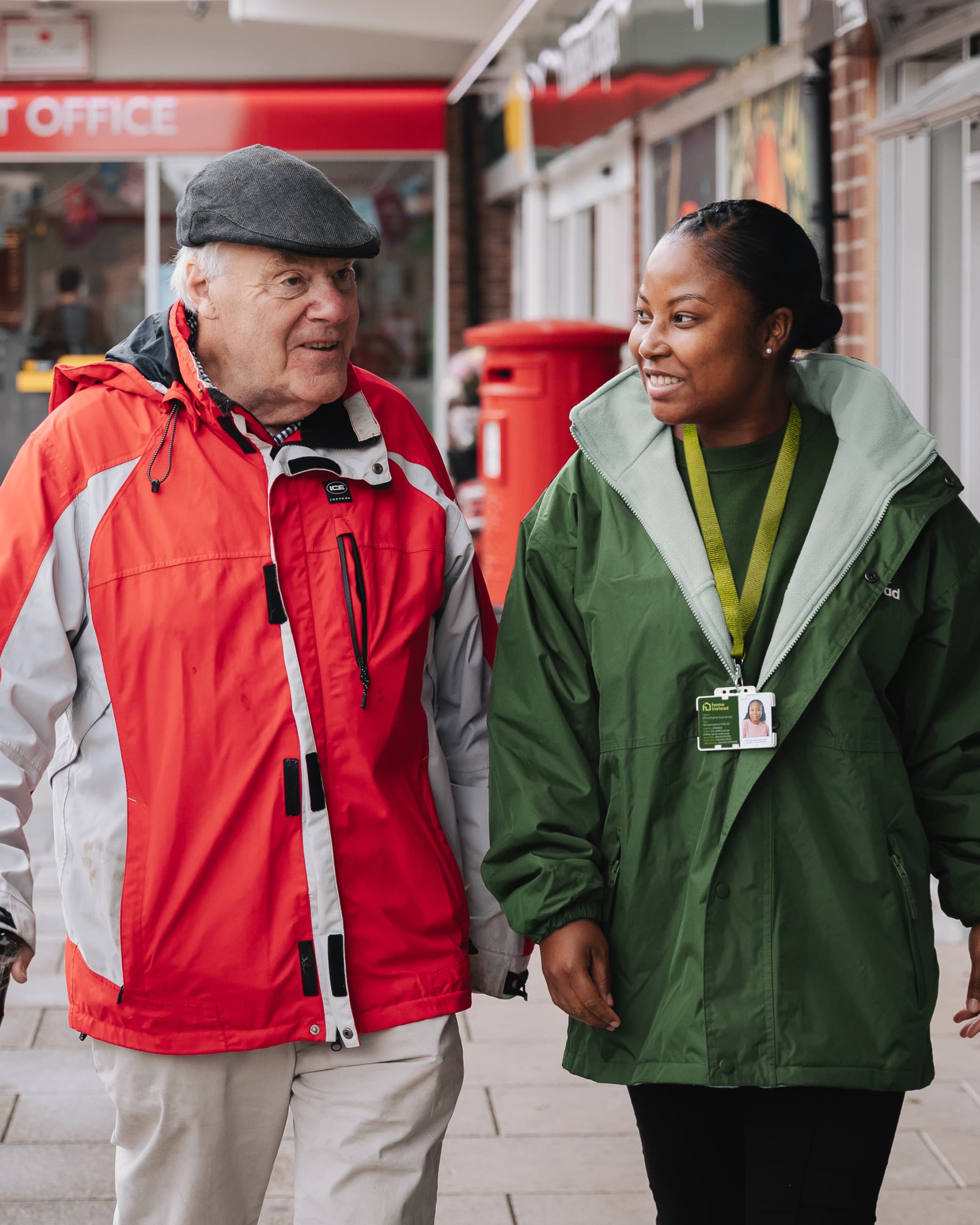 An elderly man and a young woman walk together outside, smiling and talking, both wearing jackets. - Home Instead