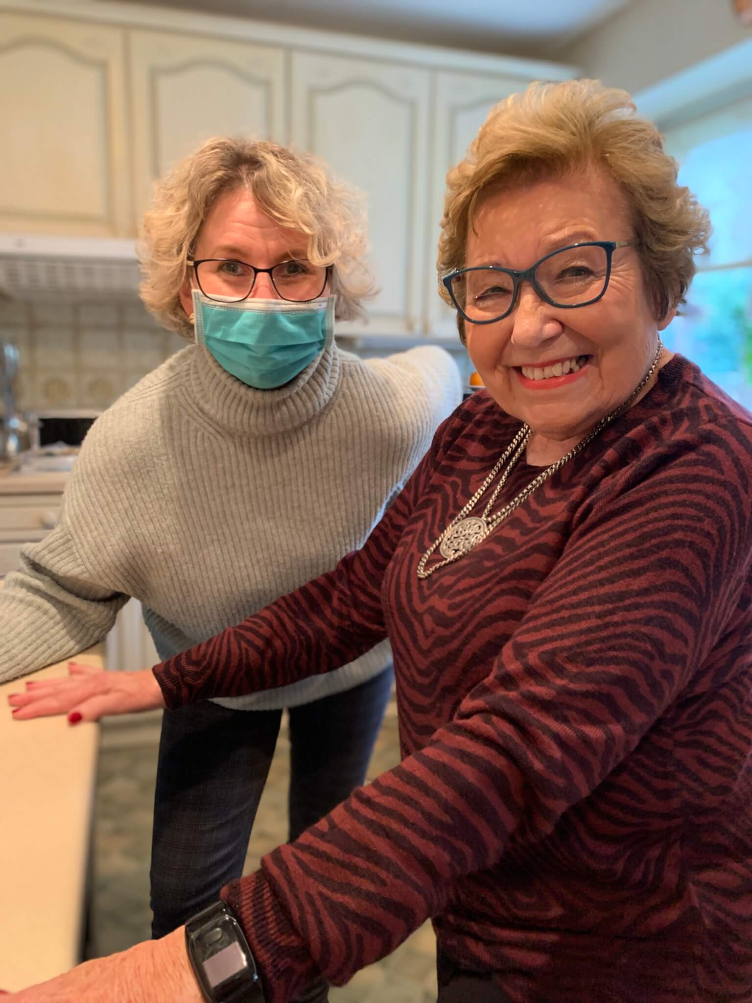 Two women smiling in a kitchen, one wearing a mask and the other in a red striped sweater with glasses. - Home Instead