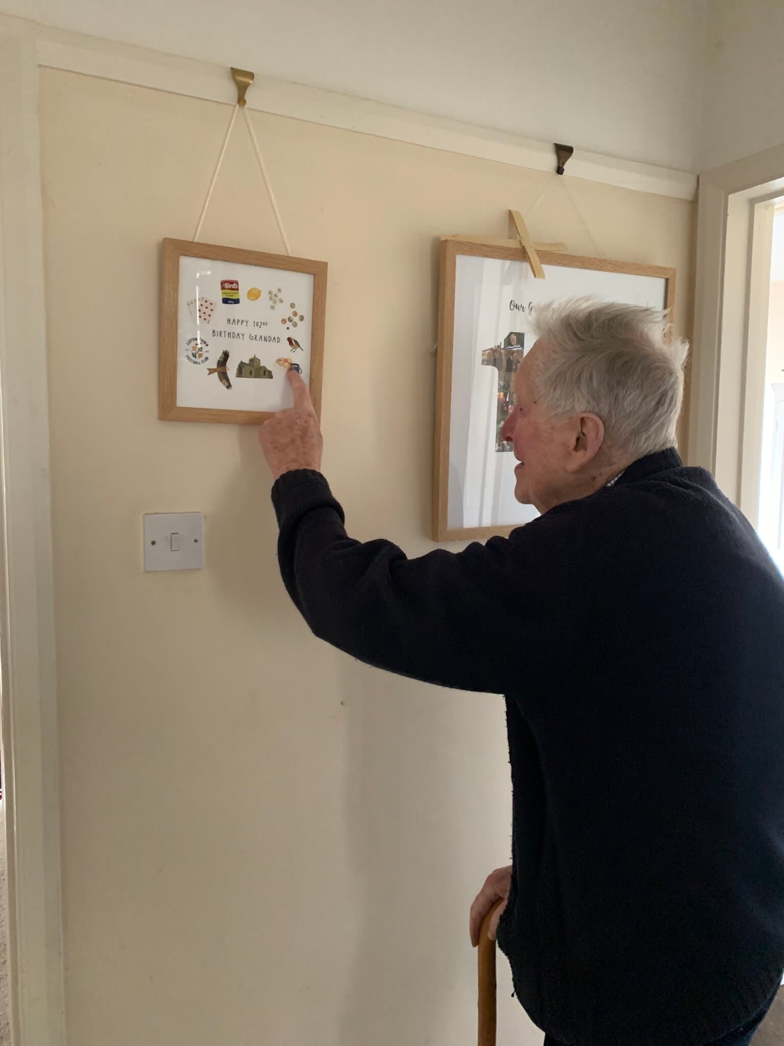 An elderly man pointing at a framed picture on the wall while standing in a hallway with the aid of a walking stick. - Home Instead