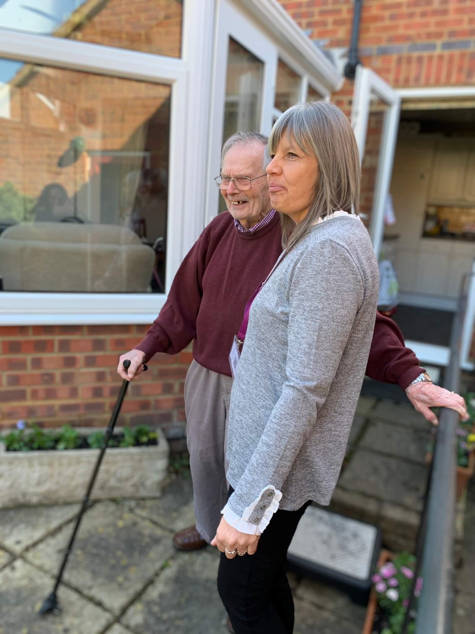 Woman standing beside an older man with a cane, smiling together outside a house with a patio in the background. - Home Instead