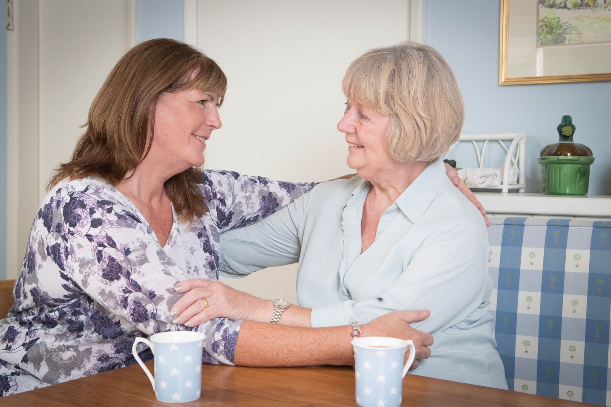 Two women, one older and one younger, smiling and embracing each other at a table with two mugs. - Home Instead