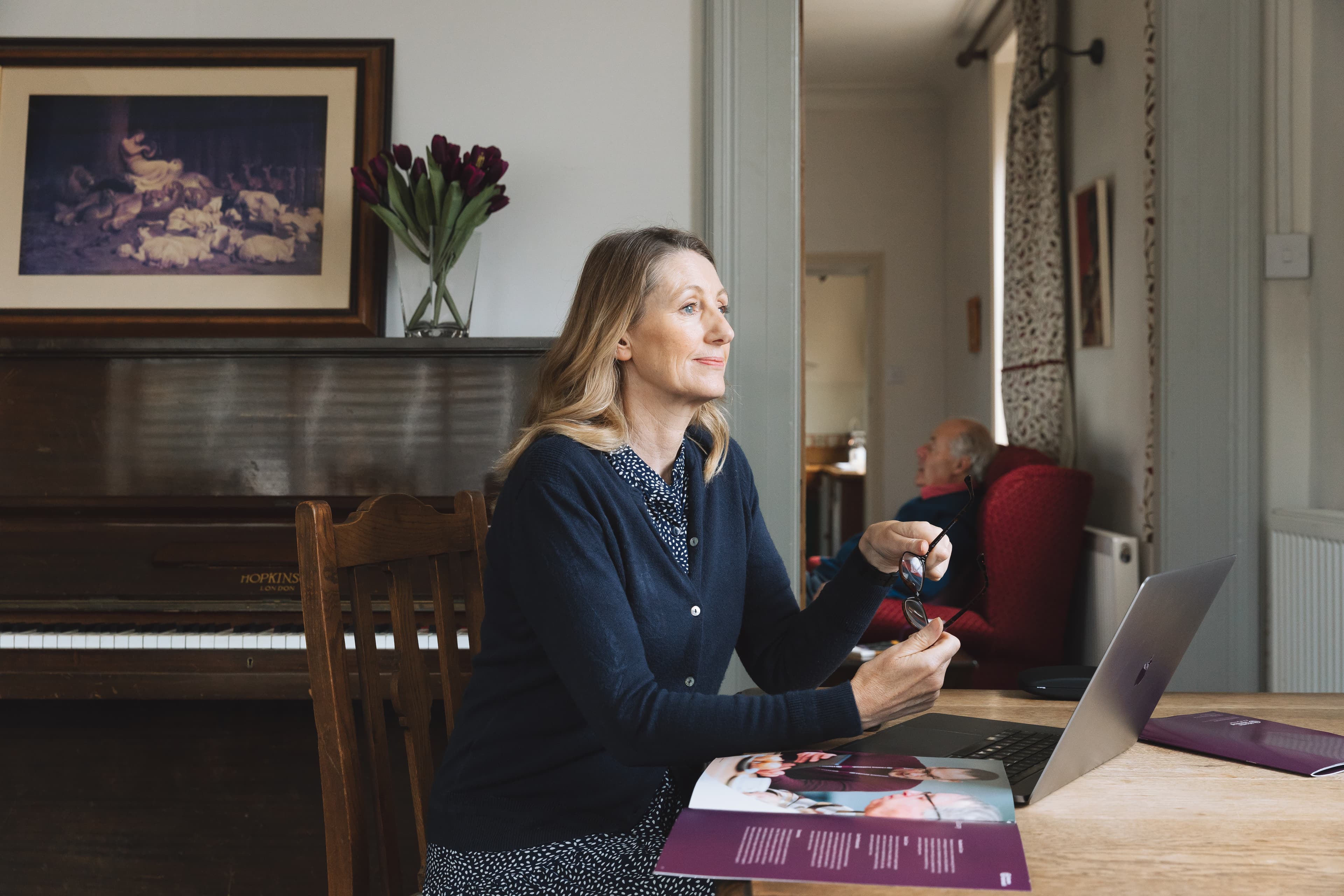 Woman sitting at a table with a laptop and magazine, holding glasses, framed painting and piano in the background. - Home Instead