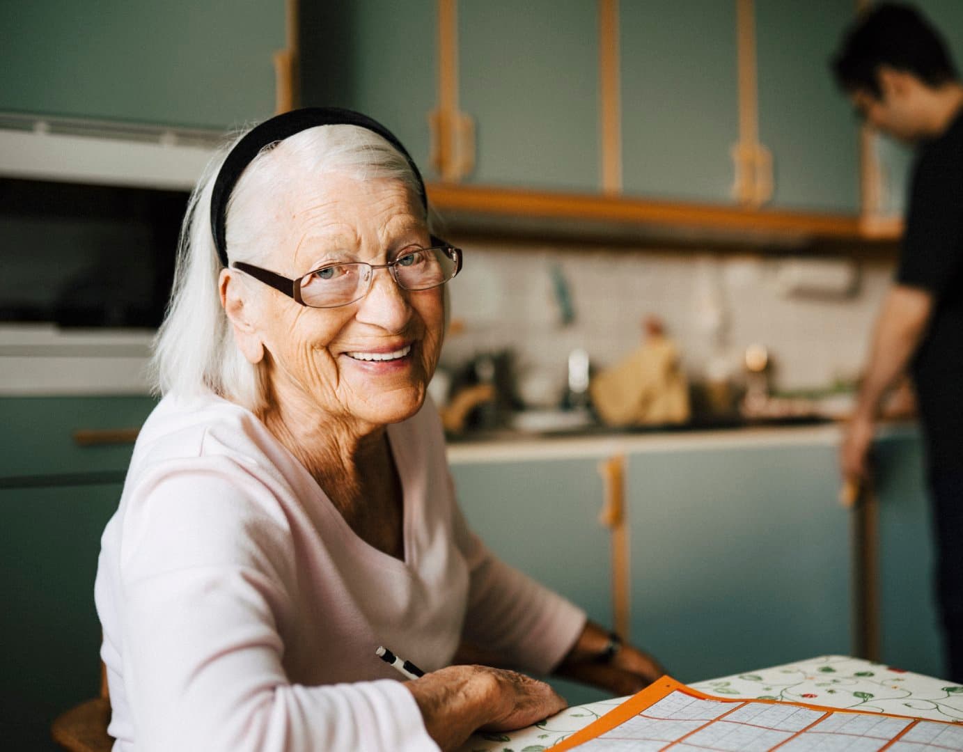 Smiling elderly woman with spectacles sits at a table with a crossword in a cosy kitchen. - Home Instead