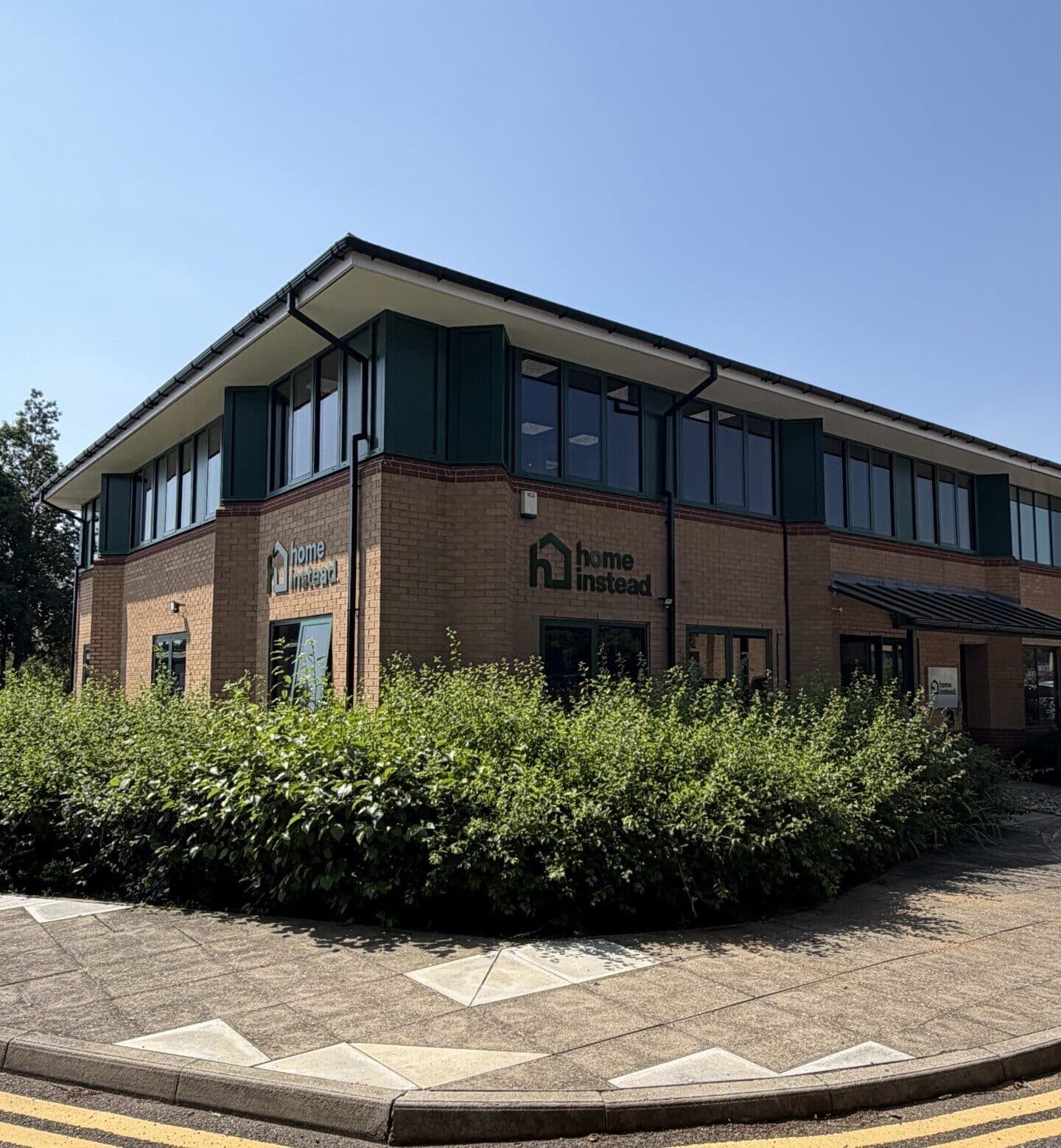 A two-storey brick office building with "Home Instead" signs and greenery in front, under a clear blue sky. - Home Instead