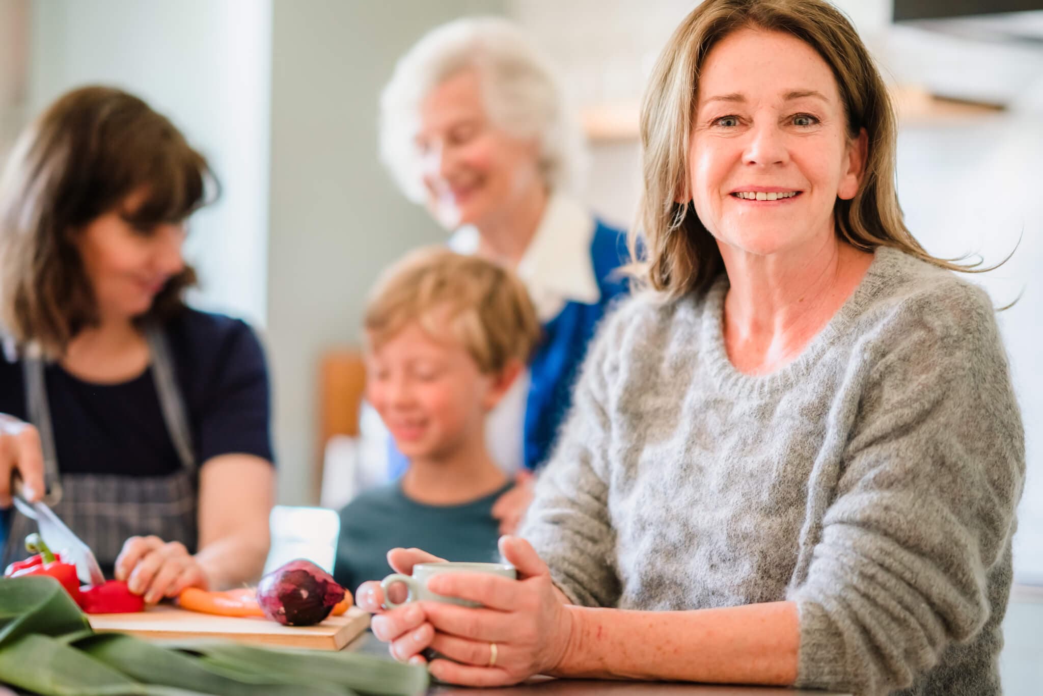 A woman smiles at the camera while others, including a child, are blurred in the background preparing food. - Home Instead