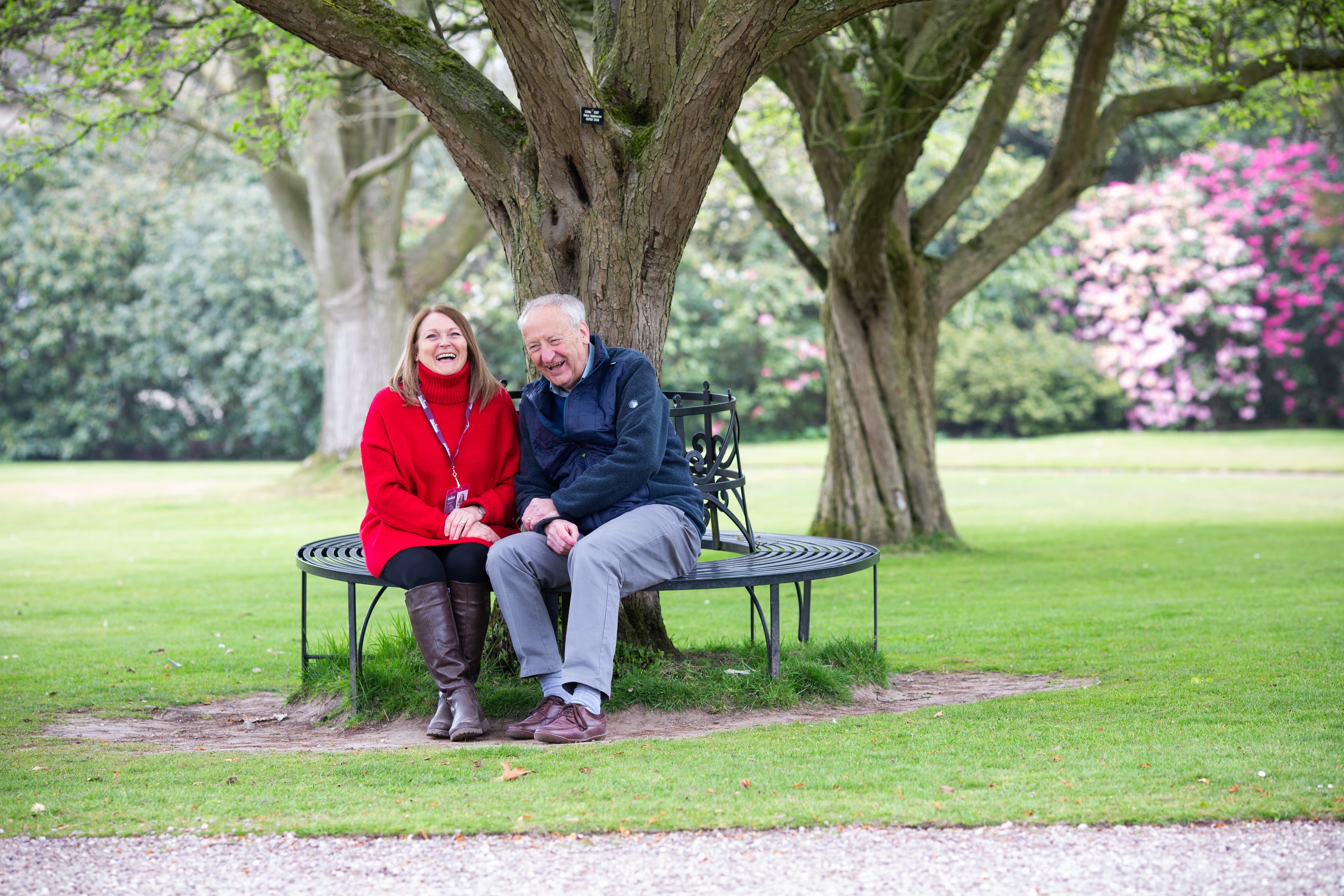 An elderly couple sitting on a curved bench under a tree, smiling and laughing in a park setting. - Home Instead