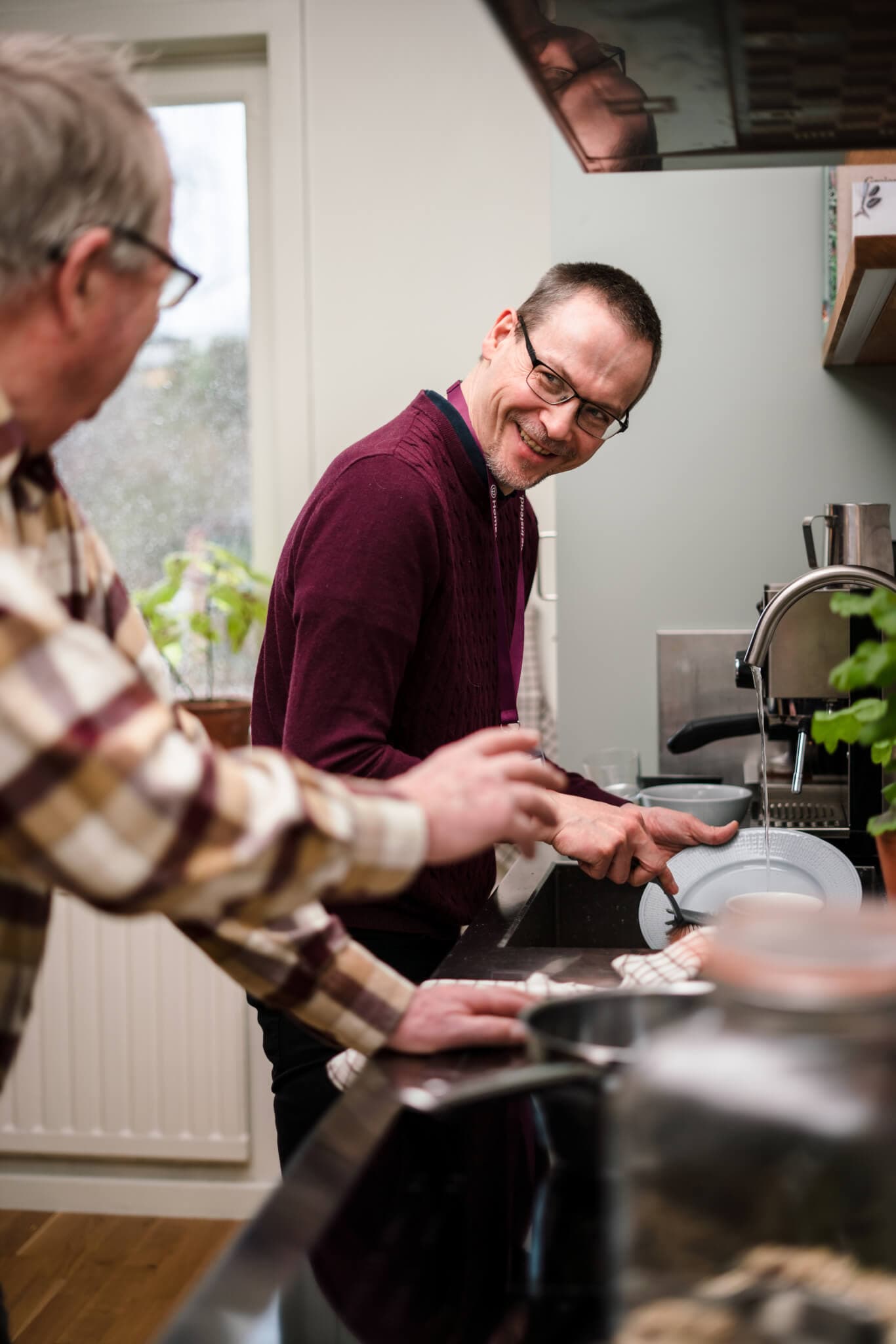 Two men, one elderly and one middle-aged, washing dishes together in a kitchen and smiling at each other. - Home Instead