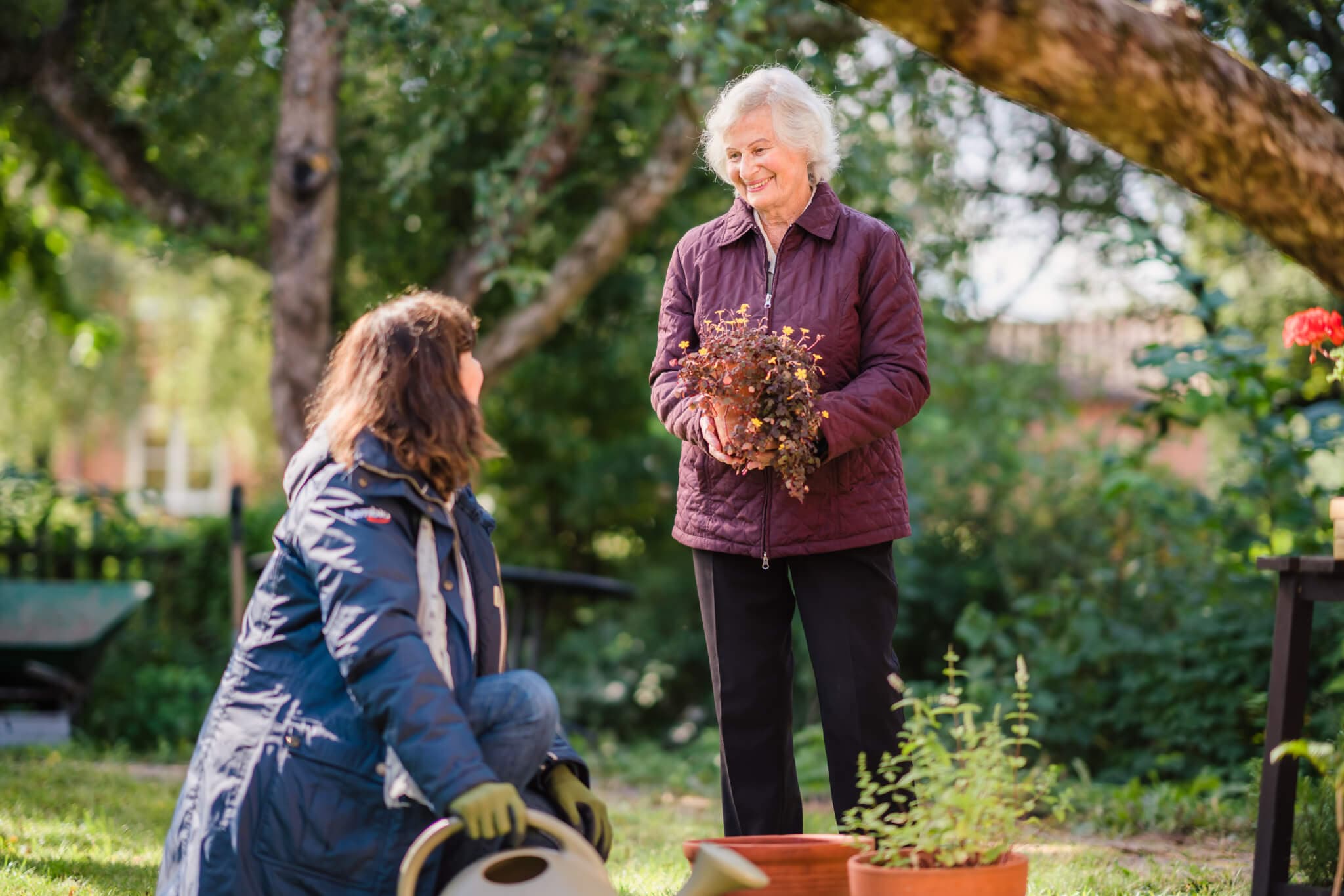 Two women gardening outdoors; one is standing holding a plant, and the other is kneeling beside pots and gardening tools. - Home Instead