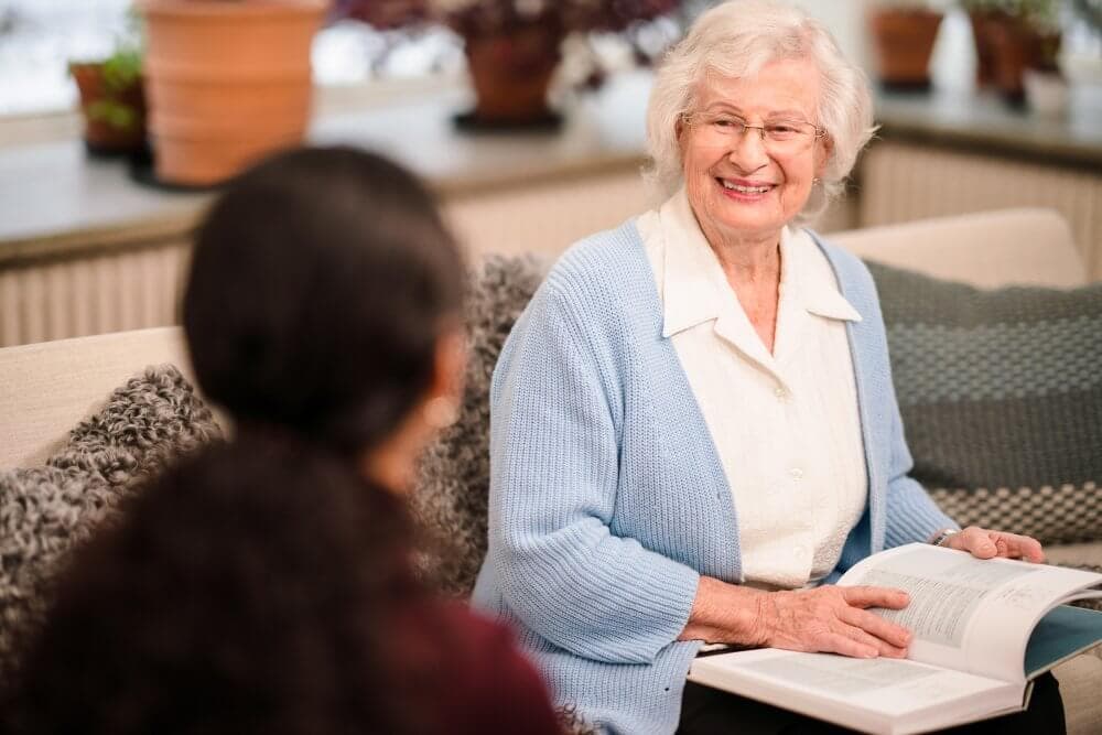 An elderly woman with glasses reading a book, smiling at a person sitting next to her in a cozy room. - Home Instead