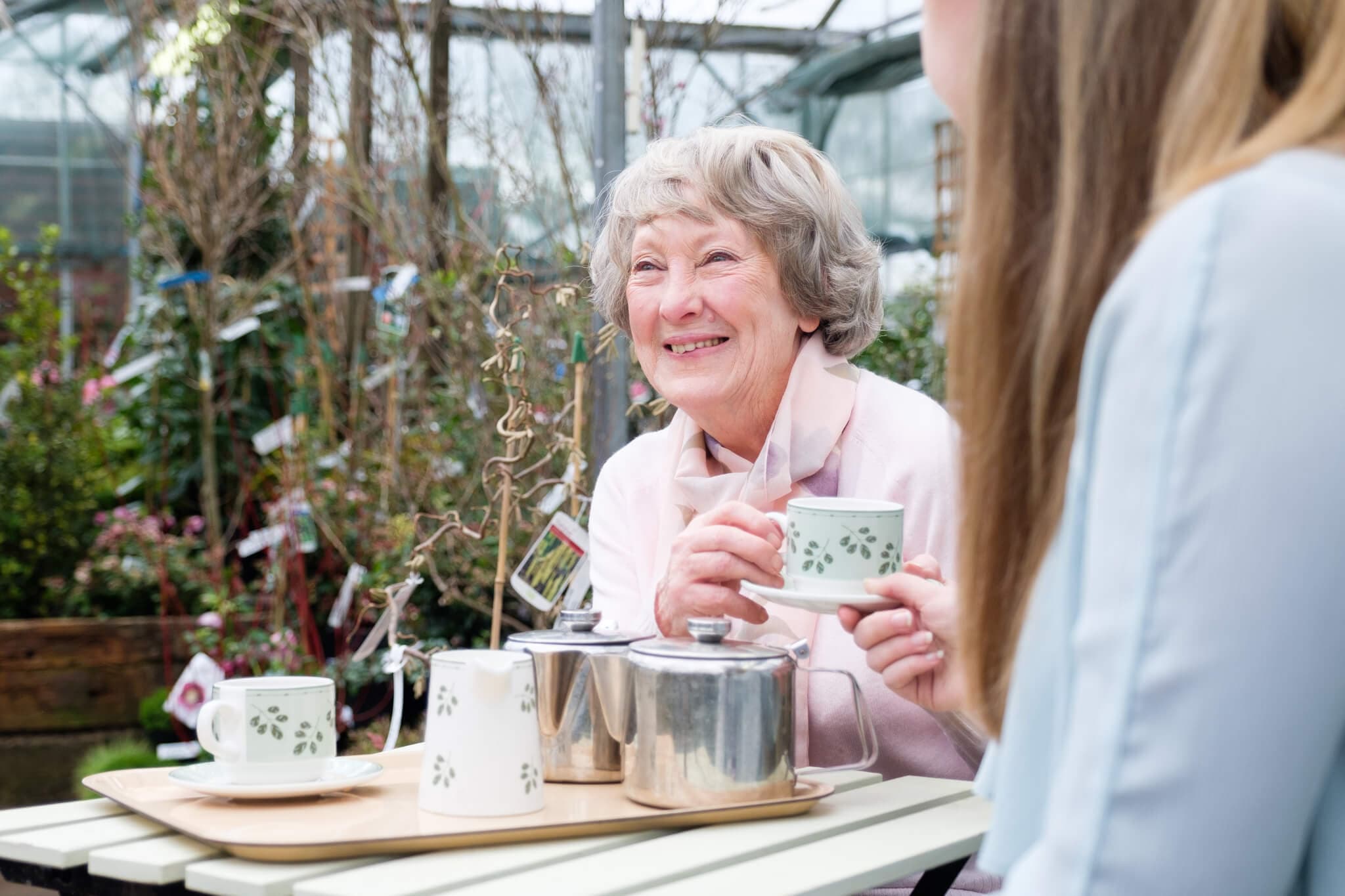 Two women, one elderly, smiling and enjoying tea together at an outdoor garden cafe. - Home Instead