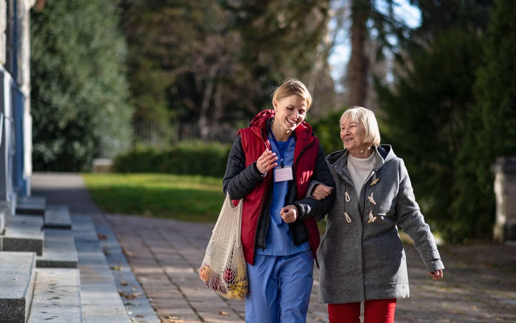 A young caregiver joyfully walks with an elderly woman on a sunny day, both engaged in conversation. - Home Instead
