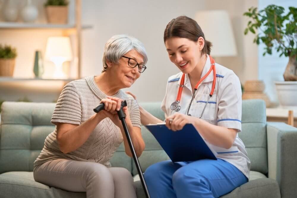 Nurse showing a clipboard to an elderly woman with a cane, while they sit on a couch in a cozy living room. - Home Instead