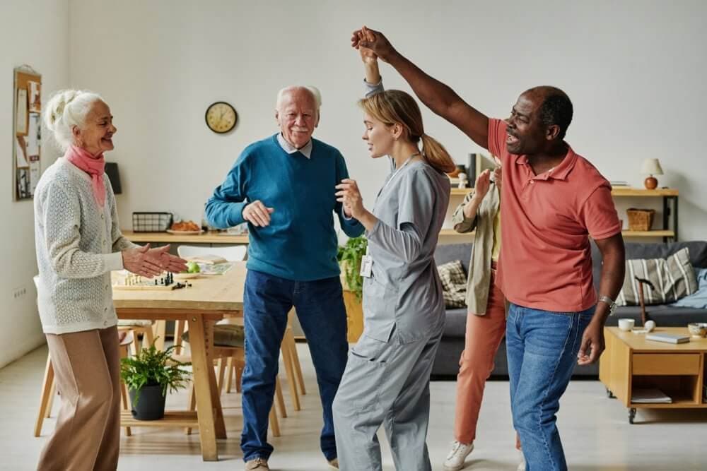 Four people, including a caregiver, dance and smile in a cozy living room with a dining table and sofa in the background. - Home Instead
