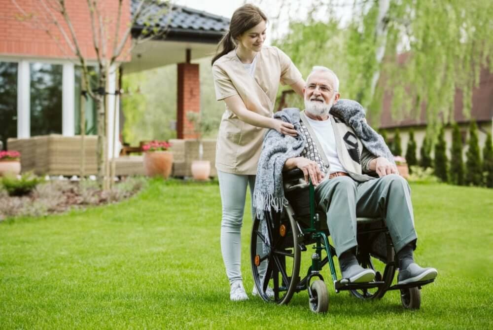 A caregiver assists an elderly man in a wheelchair outdoors in a garden area. - Home Instead