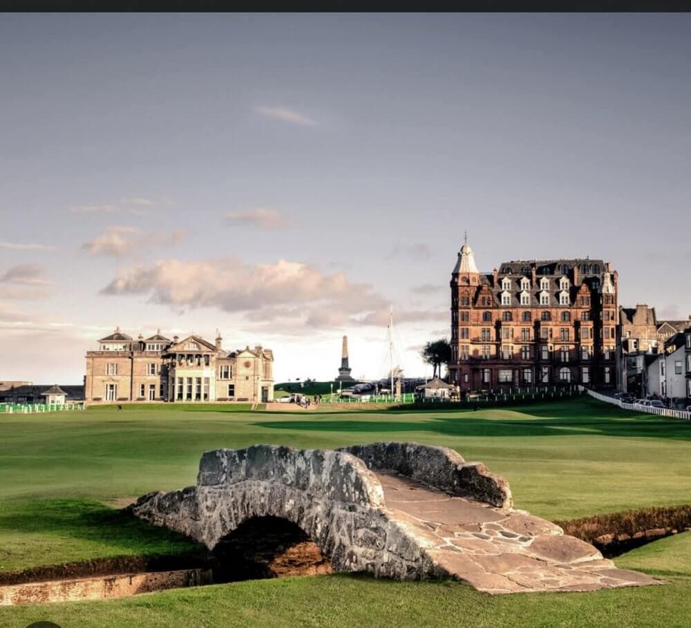 A historic stone bridge on a golf course with a large building and cloudy sky in the background. - Home Instead