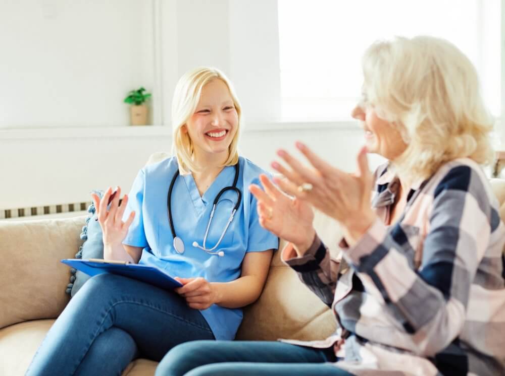 A smiling healthcare professional discussing with an elderly woman, both seated on a couch in a brightly lit room. - Home Instead