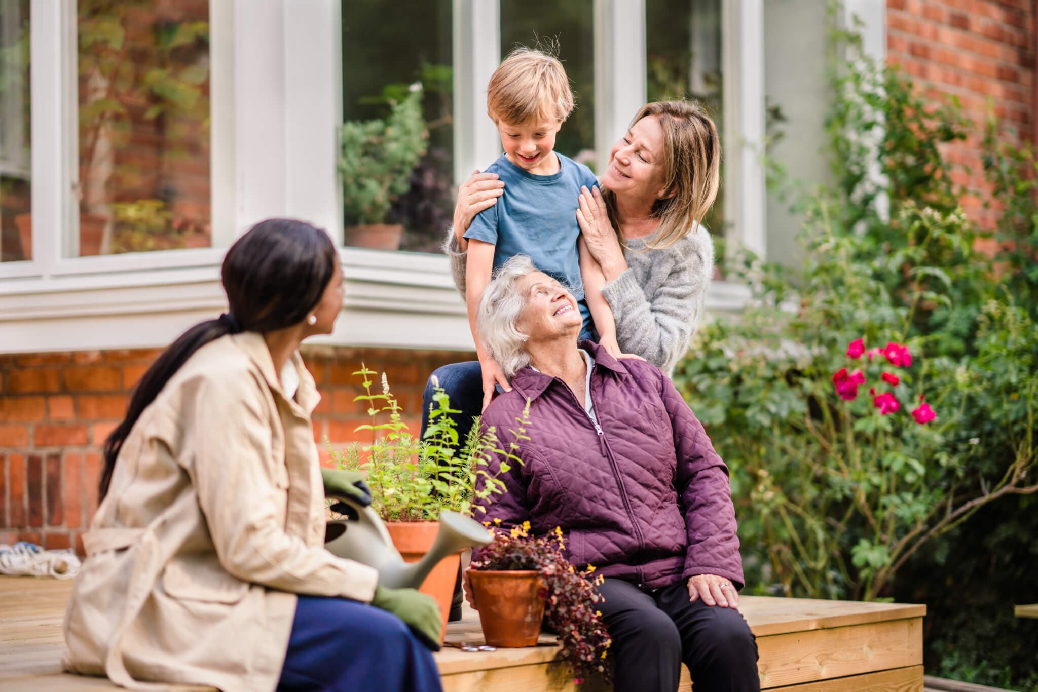 Three generations smiling: a young boy, his mom, his grandmother, and his great-grandmother in a garden. - Home Instead