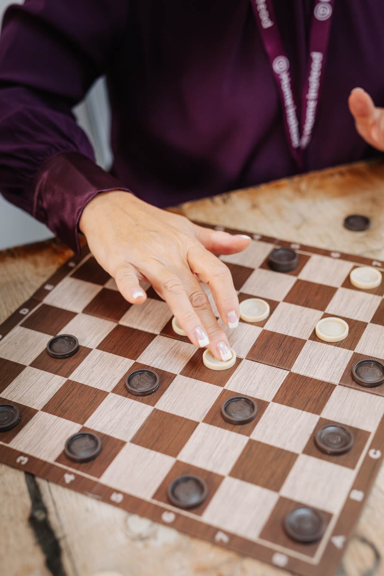 A person moves a white piece on a wooden checkerboard, surrounded by black pieces. - Home Instead