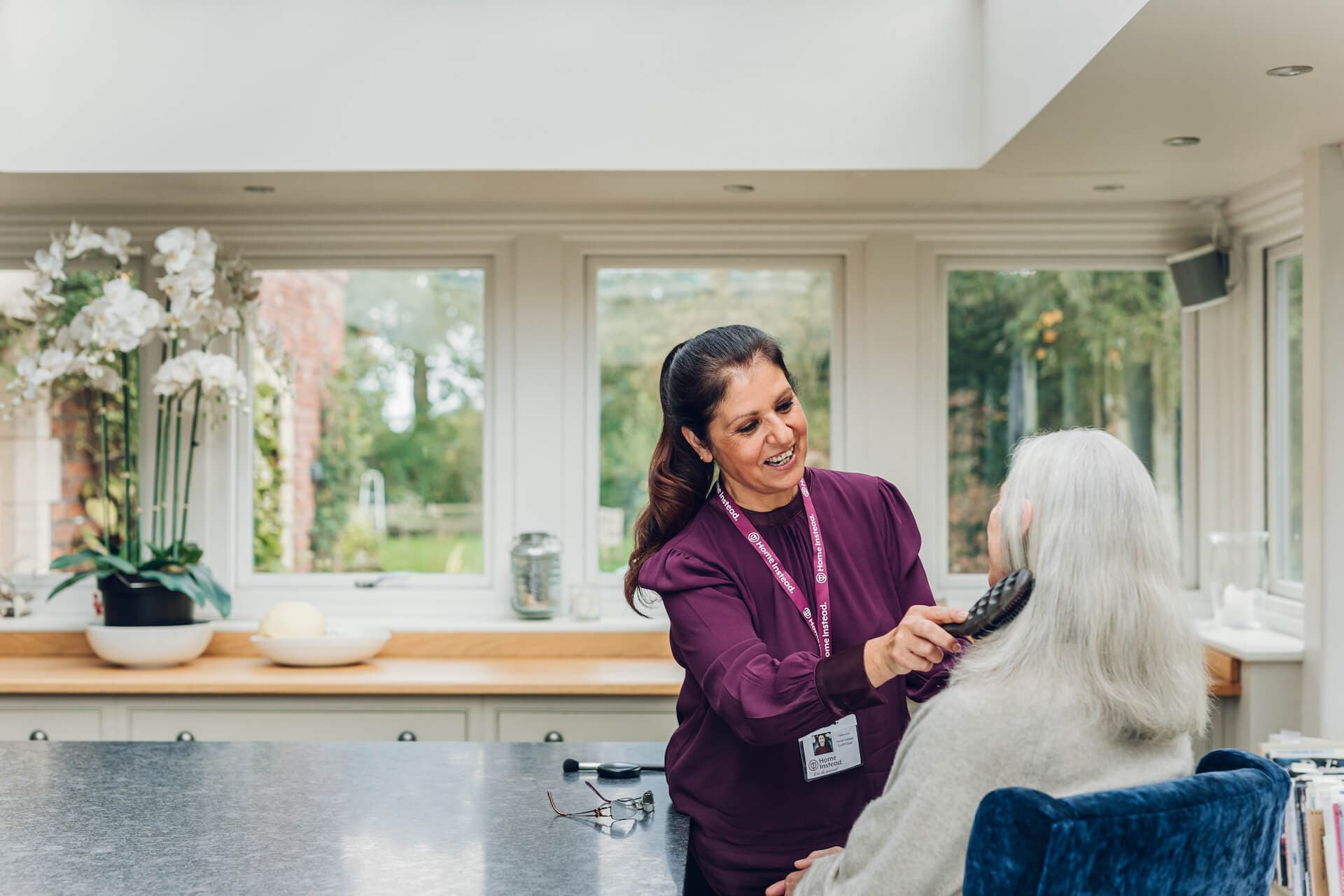 A caregiver with long dark hair, dressed in purple, brushes the white hair of an elderly person in a bright room. - Home Instead