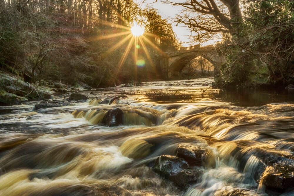 Sun setting over a tranquil river with gentle waterfalls, an arched stone bridge, and trees in the background. - Home Instead