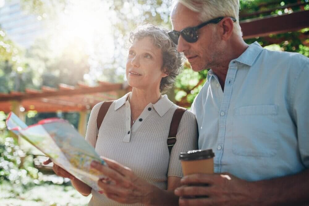 An older couple outdoors, looking at a map. The man holds a coffee cup and wears sunglasses. They appear to be navigating. - Home Instead