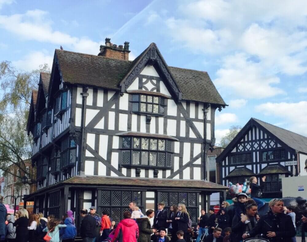 A crowd gathers in front of a black-and-white half-timbered house under a cloudy blue sky. - Home Instead