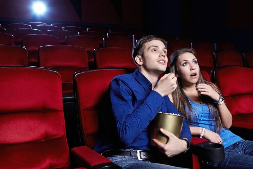 A couple sits in a theater, watching a movie with intense expressions, with red seats and an empty audience behind them. - Home Instead