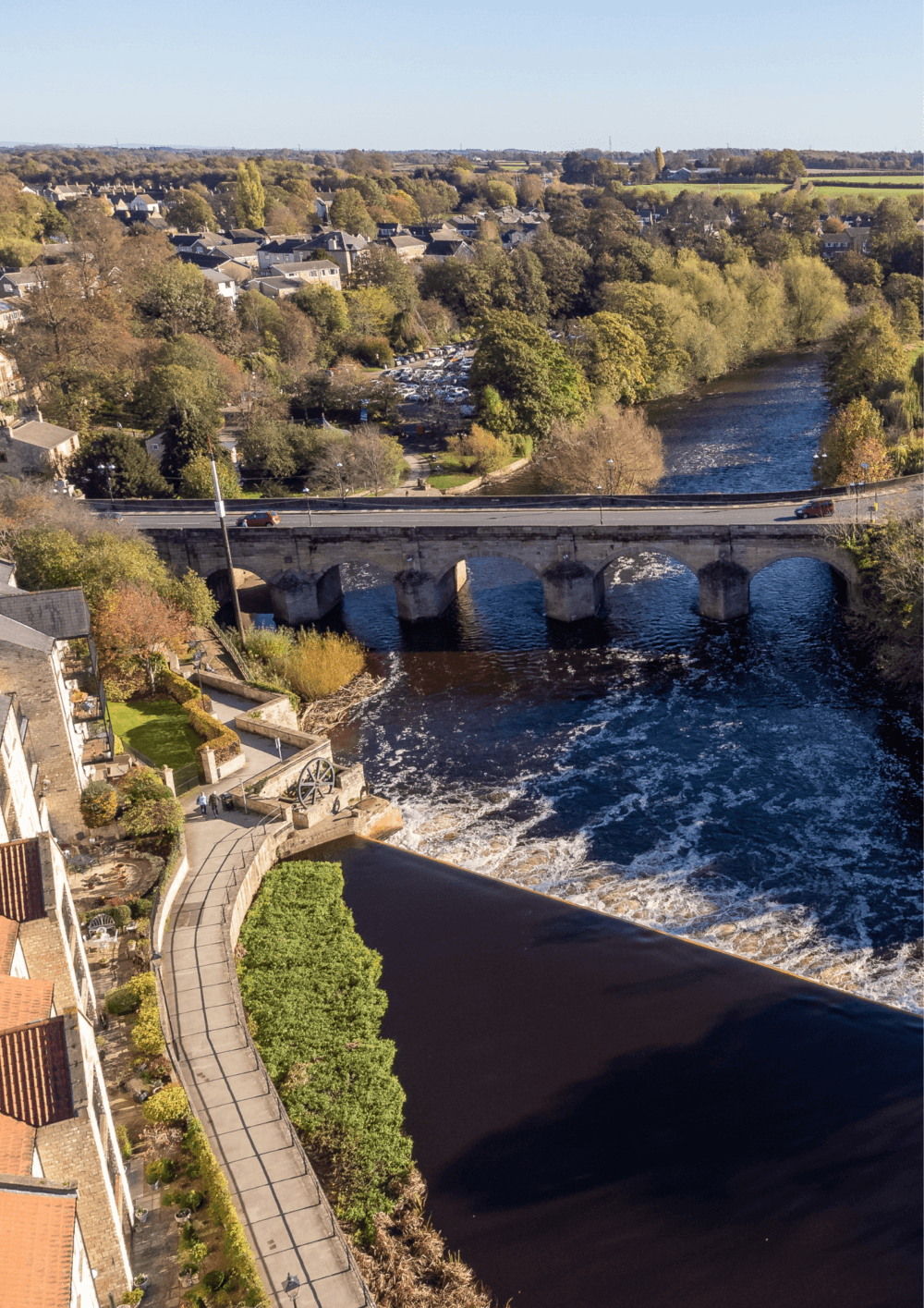 Aerial view of a bridge over a river with a park and buildings on one side and vibrant foliage around. - Home Instead