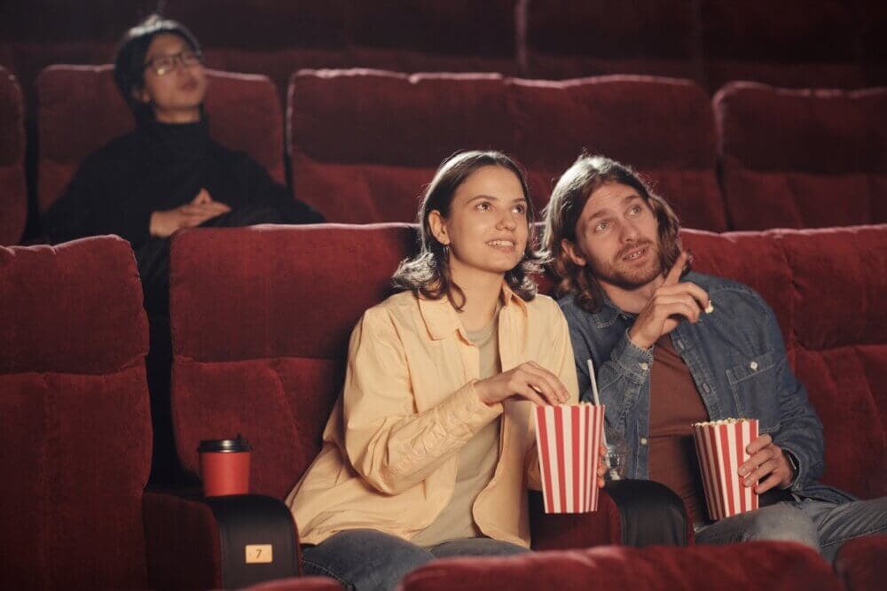 A couple enjoys popcorn while watching a movie in a nearly empty theater; another person sits several rows behind them. - Home Instead