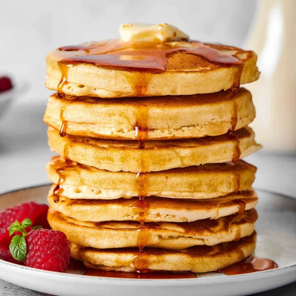A stack of pancakes topped with butter and syrup, served with fresh raspberries and mint on a plate. - Home Instead