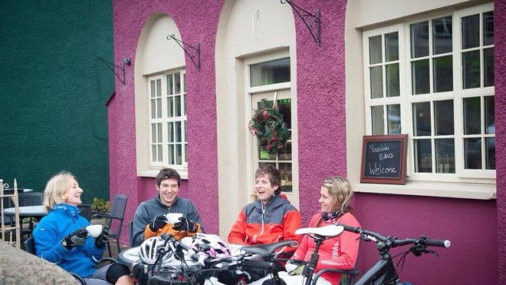 Four people in cycling gear sit and chat around a table outside a colorful cafe with their bikes parked in front. - Home Instead