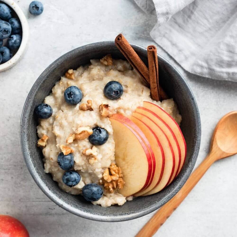 A bowl of oatmeal topped with apple slices, blueberries, walnuts, and cinnamon sticks, next to a wooden spoon. - Home Instead