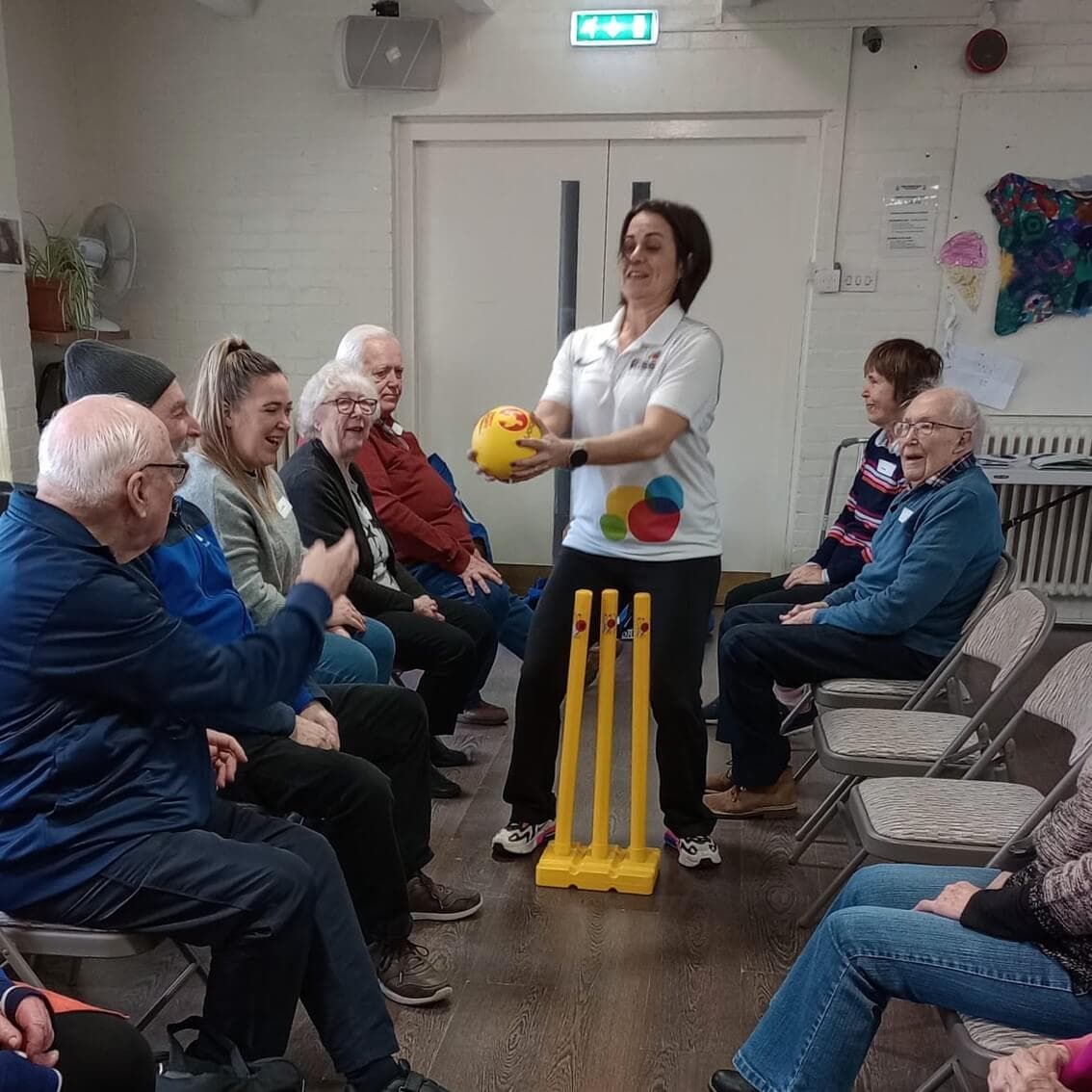 A group of elderly people sit in chairs, watching a woman holding a yellow ball near yellow plastic wickets indoors. - Home Instead