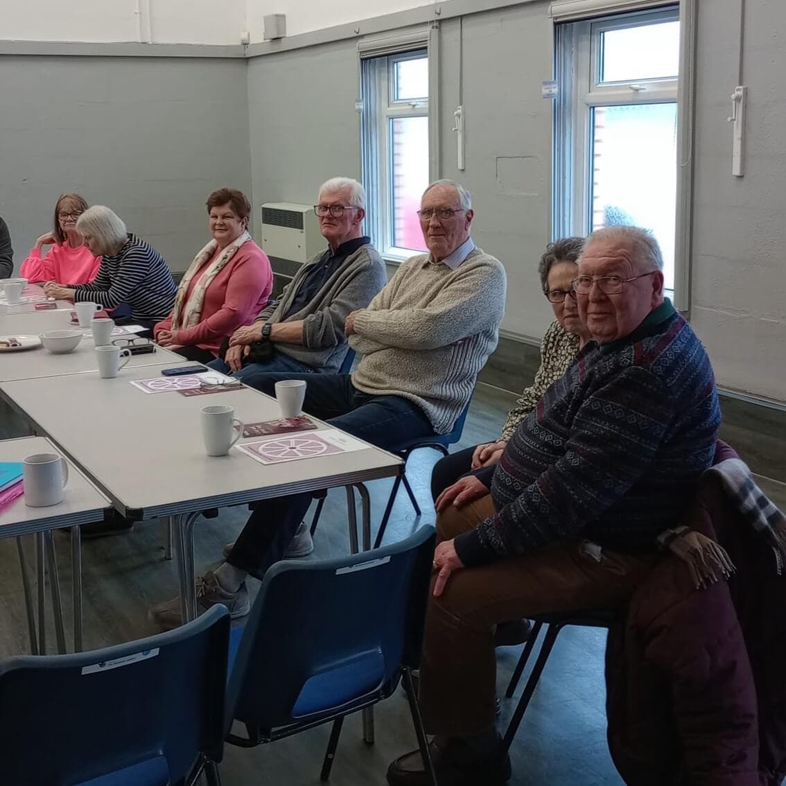 A group of seniors sit at tables in a community room, smiling, with coffee cups and pamphlets on the tables. - Home Instead