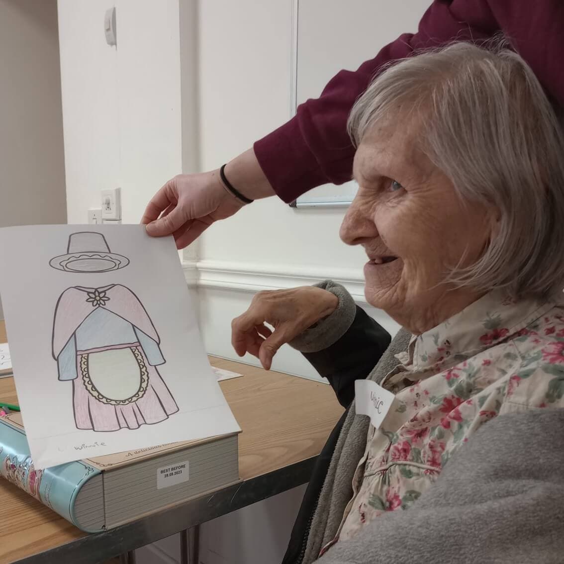 Older woman smiling, holding a drawing of a person in historic clothing with the help of another person. - Home Instead