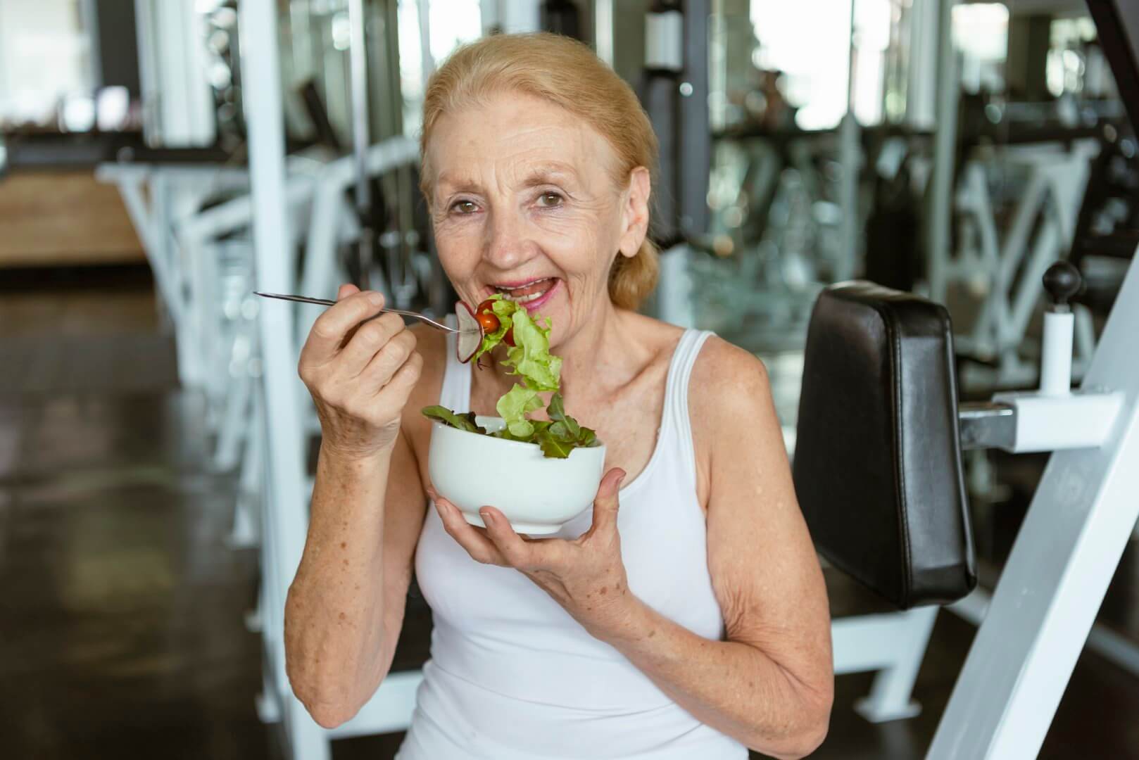 Elderly woman in a white tank top eating a salad and smiling in a gym setting. - Home Instead