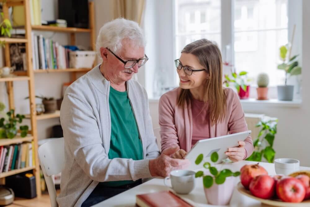 Older man and younger woman looking at a tablet together, smiling, seated at a table with books and fruits. - Home Instead Bournemouth & Christchurch