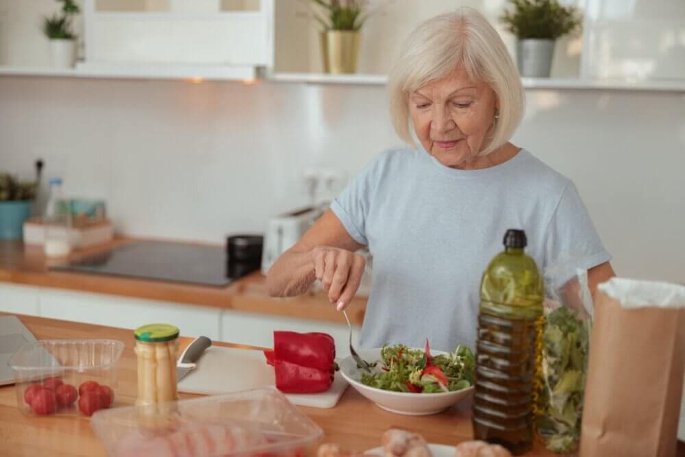 An elderly woman prepares a salad in a modern kitchen, surrounded by vegetables and cooking ingredients. - Home Instead
